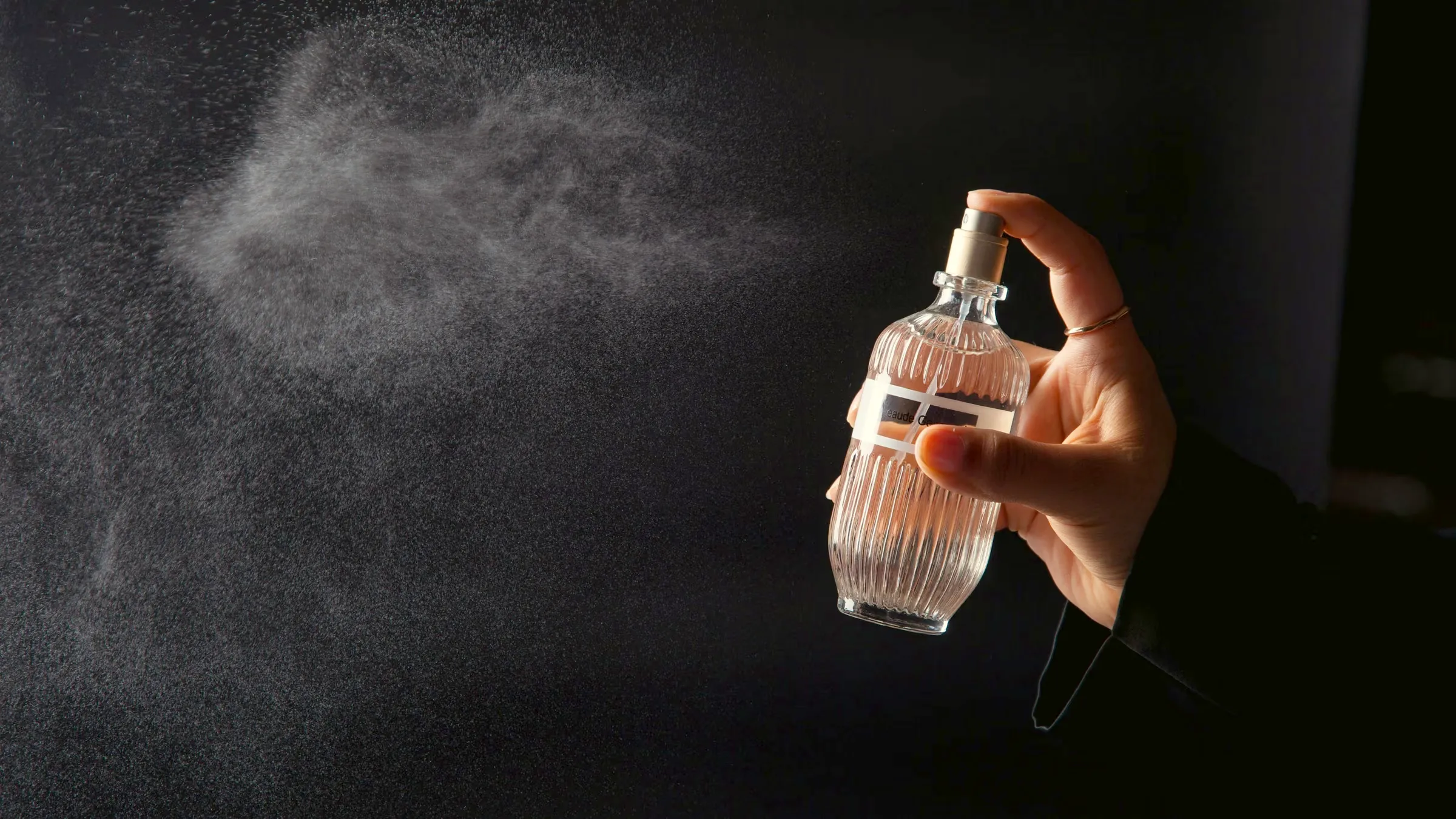 Hand misting perfume from a fluted glass bottle against a black background